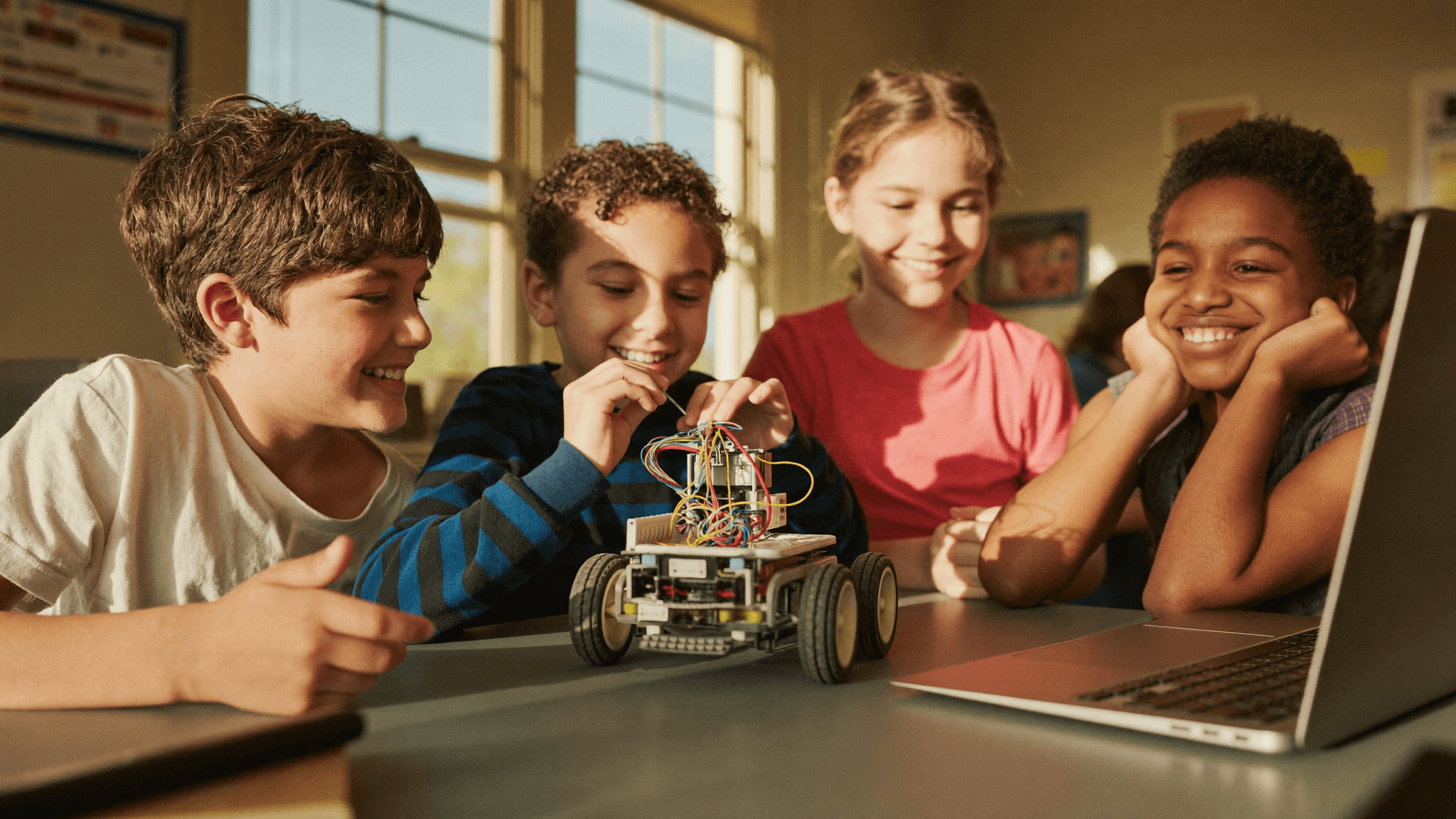 Four smiling children gathered around a small robot they built in a classroom